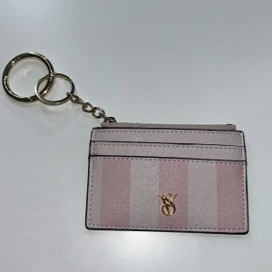 Randig rosa korthållare Victoria's Secret - Supersöt korthållare från Victoria's Secret i rosa och ljusrosa ränder med guldfärgad VS-logga framtill. Har flera kortfack, dragkedja upptill och nyckelring i metall. Perfekt att ha i väskan eller hänga på nyckelknippan. Bara använt i 2 dagar. Pris kan diskuteras.