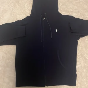 Mörkblå hoodie från Polo Ralph Lauren - Mörkblå hoodie från Polo Ralph Lauren med vit broderad logga på bröstet. Tröjan har huva med snörning, dragkedja framtill och två fickor. Tillverkad i mjuk bomull och har långa ärmar. Perfekt för en chill och stilren look.