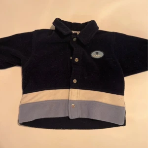 Mörkblå fleecejacka från H&M Baby - Mysig mörkblå fleecejacka från H&M Baby med krage och knappar framtill. Jackan har en beige och ljusblå rand längst ner och ett litet Baby-märke på bröstet. Perfekt för kyliga dagar och enkel att ta på.