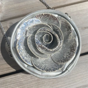 Cool silver axelväska - Säljer en rund axelväska i silver med en stor glittrig blomma framtill. Väskan har en cool metallic-look och är perfekt för dig som vill sticka ut. Väskan har dragkedja upptill! Inga defekter förekommer!😇💕