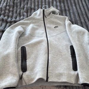 Grå Nike hoodiejacka med dragkedja - Grå hoodiejacka från Nike med svart logga på bröstet. Jackan har hel dragkedja framtill, två svarta dragkedjefickor och en hög krage. Tillverkad i mjuk bomullsblandning och har en sportig, clean look med huva och långa ärmar.