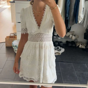 Vit spetsklänning/Studentklänning - Supersöt vit kortklänning med djup v-ringning och spetsdetaljer. Klänningen har korta ärmar med spetskant, broderade blommor och ett snyggt hålmönster i midjan. Perfekt för studenten!