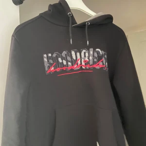 Svart hoodie från Hoodrich med tryck - Svart hoodie från Hoodrich med stor logga i vitt och rött framtill och baktill. Tröjan har huva med snörning och en stor magficka. Mjuk insida och klassisk passform, perfekt för chill eller streetwear. Snygga detaljer med texten 'From nothing to something' nedtill.