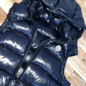 Moncler väst - Moncler Bormer väst – klassisk och tidlös Snygg och exklusiv dunväst från Moncler i modellen Bormer. Väst med lätt vaddering av högkvalitativt gåsdun som håller dig varm samtidigt som den är smidig och lätt. Klassisk design med hög krage.  Perfekt att bära över en hoodie, tröja eller skjorta – både till vardags och mer uppklätt. Ger en sportig men samtidigt stilren look som är typisk för Moncler. 	•	Storlek L