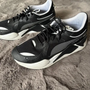 Svarta och vita sneakers från Puma - Snygga svarta och vita sneakers från Puma med coola detaljer och kontrasterande mönster längs sidorna. Skorna har snörning, rund tå och en chunky platt sula. Materialmix av syntet och mesh ger både stil och komfort. Perfekta för dig som gillar sportig streetstyle.