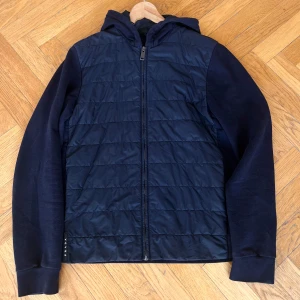 Mörkblå cardigan från Prada - Clean och simpel mörkblå cardigan från Prada Cardiganen har huva, dragkedja och diskret Prada-logga vid nederkanten. Perfekt för dig som vill ha en stilren höstjacka! Storlek: S