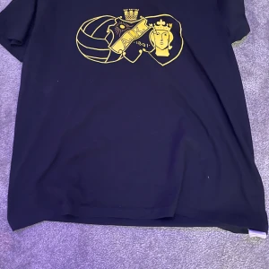 Svart AIK t-shirt med gult tryck - Svart t-shirt från Södra tifo i storlek S med ett stort gult tryck framtill som visar klubbens emblem och detaljer. Klassisk passform och rund halsringning, perfekt för dig som vill visa ditt AIK-stöd. Gjord i mjukt bomullstyg. Lite stor i halsen 