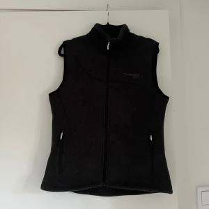 Mörkgrå fleeceväst från Rosetti  - Mysig mörkgrå fleeceväst från Rosetti Riding med hög krage och hel dragkedja framtill. Västen har två praktiska fickor med dragkedja och diskret broderad logga på bröstet. Har använts i stall, enstaka hår kan därför förekomma. Färgen är mörkgrå, se sista bilen för tydligare färg.