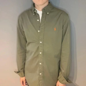 Olivgrön skjorta från Ralph Lauren… prutbar!! - Säljer en olivgrön skjorta från Ralph Lauren med klassisk button-down krage och orange broderad logga på bröstet. Skjortan har långa ärmar, vita knappar och är tillverkad i mjuk bomull. Perfekt för en clean och stilren look.          !!!prut bart!!!