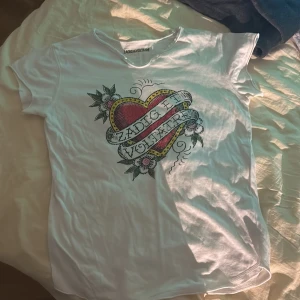 Zadig & Voltaire T-shirt - Vit t-shirt från Zadig & Voltaire med stort färgglatt hjärtmotiv och blommor på bröstet. Klassisk rund halsringning och korta ärmar. Trycket har coola detaljer och text. Hittar ingen storlek men är som en XS/S. Nypris 900kr men säljs inte längre