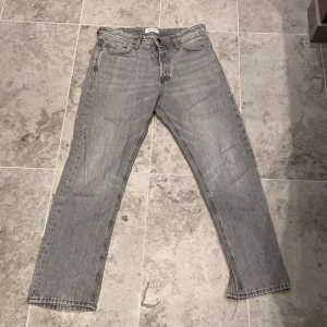 Grå Jack Jones jeans straight fit - Säljer ett par klassiska gråa Jack Jones jeans med rak passform. Jeansen har fem fickor, knappgylf och den ikoniska läderpatchen bak i midjan. Perfekt för dig som gillar en tidlös och avslappnad look.