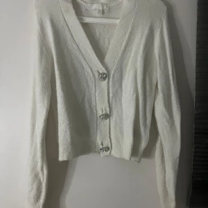 Vit kofta med glittriga knappar H&M - Superfin vit kofta från H&M med långa ärmar och snygga glittriga knappar framtill. Koftan är stickad och har en ribbad kant vid ärmslut och nederkant. Perfekt för lager-på-lager och ger en clean look med lite extra bling.