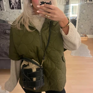 Olivgrön quiltad poncho  i storlek S - Säljer en olivgrön quiltad poncho med halv dragkedja framtill. Västen har en loose passform och är perfekt att slänga över en stickad tröja. Materialet är syntet och dun, vilket gör den både lätt och varm. Enkel och stilren design utan synliga loggor.