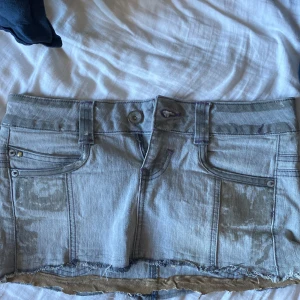 Grå jeanskjol från Resha Denim - Säljer en grå jeanskjol från Resha Denim med rå kant nertill och klassiska fickor fram och bak. Kjolen har bälteshällor, knapp- och dragkedjestängning samt lila sömmar som cool detalj. Perfekt för dig som gillar en edgy och avslappnad stil.