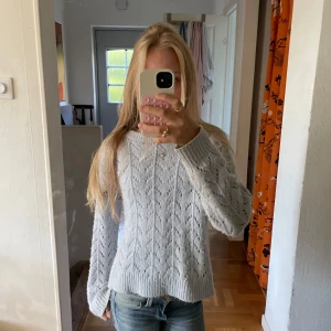 Ljusgrå stickad tröja med mönster - Superfin ljusgrå stickad tröja med snyggt hålmönster och ribbade muddar vid ärmslut och nederkant. Tröjan har långärmad passform och är perfekt för att hålla sig varm och ändå se chill ut. Passar till jeans eller kjol för en casual vibe.