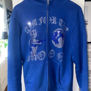 Blå hoodie Aim For The Moon L - Blå hoodie med dragkedja från Aim For The Moon. Framsidan har ett stort tryck med glittriga detaljer och text, samt en cool figur och jordglob. Tröjan har huva, fickor och långa ärmar. Perfekt för dig som gillar streetwear och vill sticka ut.