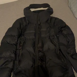 Svart dunjacka från Canada Goose - Säljer en svart dunjacka från Canada Goose med hög krage och huva. Jackan har flera dragkedjor, två sidofickor och den klassiska Canada Goose-loggan på bröstet. Perfekt för kalla dagar och riktigt snygg streetstil. Obs dragkjedan e sönder men de går att fixa 