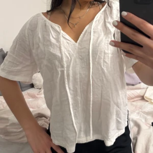 Vit blus med v-ringning - Säljer en luftig vit blus med v-ringning och knytband framtill. Blusen har korta ärmar och en avslappnad passform, perfekt för varma dagar. Enkel och stilren design som är lätt att matcha med olika outfits.
