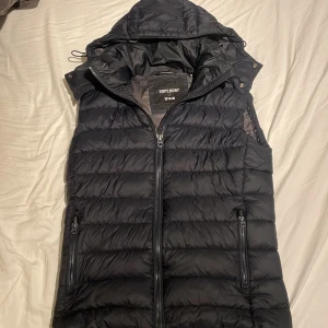 Mötkblå dunväst med huva från Superdry - Mörkblå dunväst från Superdry med avtagbar huva och två dragkedjefickor framtill. Västen är quiltad och har en hög krage samt hel dragkedja. Riktigt bra skick och sitter bra🌟