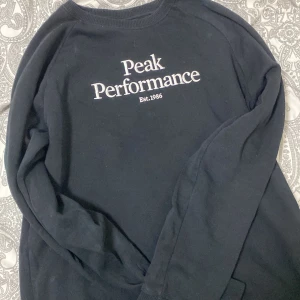 Svart Peak Performance sweatshirt - Svart sweatshirt från Peak Performance med broderad vit logga och text på bröstet. Klassisk rund halsringning och långa ärmar. Mjuk och skön bomullsblandning, perfekt för chill eller skola.