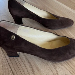 Bruna mockapumps med låg klack - Snygga bruna pumps i mocka med låg klack och rund tå. Skorna har en klassisk form och en liten metallknapp med dekorativ detalj på sidan. Perfekta för dig som gillar en stilren och tidlös look.