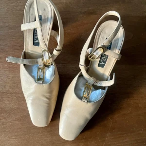 Beige klackskor från Nouchka - Snygga beige klackskor från Nouchka med fyrkantig tå och bred klack. Skorna har justerbara remmar med guldfärgade spännen och dekorativt N-detalj framtill. Tillverkade i skinn för en lyxig känsla och passar perfekt till dig som gillar stilrena och trendiga skor.