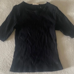 Svart ribbad topp med halvärm - En svart ribbad topp med rund halsringning och halvärm. Toppen har en enkel och stilren design med diskret logga nertill. Perfekt för att matcha med jeans eller kjol för en clean look.