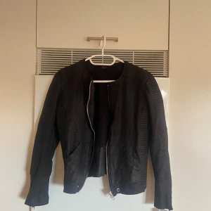 Svart bikerinspirerad jacka S - Svart jacka i bikerstil med markerade axlar och ribbade detaljer längs ärmarna. Jackan har dragkedja framtill och vid ärmsluten, samt två fickor. Snygg och stilren modell som passar till många outfits.