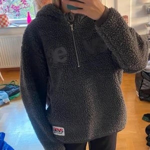 Grå teddy hoodie från Svea - Mysig grå teddy hoodie från Svea med broderad logga på bröstet och patch nertill. Tröjan har huva med dragsko, half zip och är tillverkad i fluffig fleece. Perfekt för kalla dagar och chill vibes.