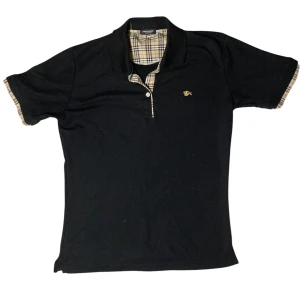 Svart Burberry piké med rutiga detaljer - Svart piké från Burberry med klassiska beigerutiga detaljer i krage och ärmslut. Liten guldfärgad brodyr på bröstet och knappslå framtill. Snygg och tidlös design som sticker ut med de ikoniska rutorna. Det är storlek XXL i junior så passar mellan 160 till 170 cm lång. Tveka inte på att komma med frågor eller prisförslag ⬛️⚜️