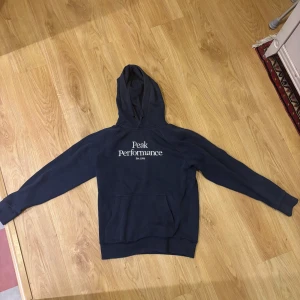 Mörkblå hoodie från Peak Performance - Snygg mörkblå hoodie från Peak Performance med vit logga på bröstet. Klassisk känguruficka framtill, huva med snörning och långa ärmar. Perfekt för chill dagar eller när du vill ha en stilren look.