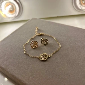 Guldigt smyckeset med rosor - Snyggt smyckeset med armband och örhängen i guldton. Båda delarna har en öppen rosdesign som ger en romantisk och trendig vibe. Kedjan är tunn och smidig, perfekt för att lyfta din stil med en subtil men iögonfallande detalj.