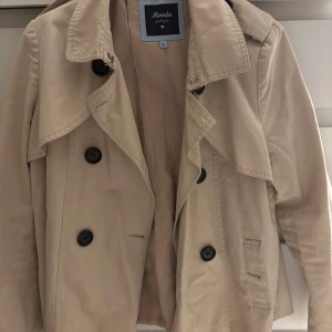 Snygg trenchcoat  - Stilren beige trenchcoat från Moeda med klassisk dubbelknäppning, bred krage och snygga axelremmar med knappar. Jackan har fickor framtill och är tillverkad i ett mjukt bomullsmaterial. Perfekt för dig som vill ha en tidlös look.