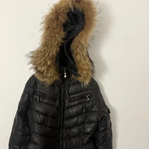 Hollies jacka  - Pris kan diskuteras nästan ny bättre bilder kan skickas privat org pis 3500kr Svart pufferjacka från Hollies med stor huva och brun fuskpäls. Jackan har dragkedja framtill, två bröstfickor med dragkedja och bälte i midjan. Insidan har ett snyggt spetsmönstrat foder. Perfekt för kalla dagar.