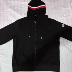 Svart Moncler zip - Svart hoodie från Moncler med hel dragkedja, huva och snörning. Röd och vit rand på huvan samt muddar i rött och vitt. Klassisk Moncler-logga på ärmen. Perfekt för dig som gillar stilrena streetwear-plagg.