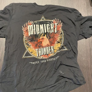 Svart t-shirt Midnight Thunder - Cool svart t-shirt med stort tryck på framsidan: en örn med röda och orange vingar, texten 'MIDNIGHT THUNDER' och 'Faster Than Heartbeats'. Rund hals och korta ärmar, perfekt för dig som gillar rockiga motiv.