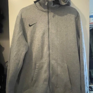 Grå hoodie från Nike med dragkedja - Grå Nike hoodie i storlek L med hel dragkedja framtill och huva. Tröjan har två stora fickor och ett svart Nike-logga på bröstet. Tillverkad i mjuk bomullsmix, perfekt för chill eller träning. Klassisk och enkel design med lång ärm och normal passform.