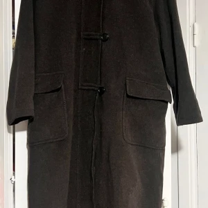 Brun ullrock från Erich Fend, strl 42 - Snygg brun rock från Erich Fend i storlek 42. Tillverkad i mjuk ullmix med klassisk krage, stora fickor och knappar framtill. Fodrad med slätt tyg och har en diskret dragkedja under knappslån. Perfekt för dig som gillar stilrena och tidlösa plagg.