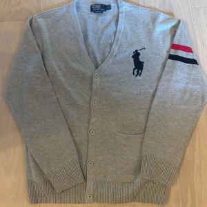 Grå kofta från Polo Ralph Lauren XL - Grå kofta från Polo Ralph Lauren i klassisk fit. Stort mörkblått broderat polospelare-logo på bröstet och coola ränder i rött, vitt och blått på ena ärmen. Koftan har knappar framtill, ribbade muddar och en ficka. Perfekt för lager-på-lager-stil.
