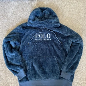 Blå fleece hoodie Polo Ralph Lauren - Supermjuk och fluffig mörkblå hoodie från Polo Ralph Lauren med stor broderad logga på bröstet och klassisk huva med snörning. Tröjan har känguruficka och ribbade muddar vid ärmslut och nederkant. Perfekt för kalla dagar när du vill vara både varm och snygg. 
