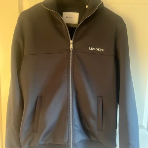 Hel zip Les Deux tröja - Snygg svart bomberjacka från Les Deux med stilren design och diskret logga på bröstet. Jackan har dragkedja framtill, två fickor och ribbade muddar vid ärmslut och midja. Perfekt för dig som gillar en clean och modern look. Sitter bra på mig som är 183