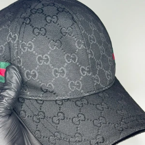 Svart keps från Gucci med GG-mönster - Snygg svart keps från Gucci med klassiskt GG-monogram över hela kepsen och en detalj i grönt och rött på sidan. Kepsen har böjd skärm och är tillverkad i slitstarkt material, perfekt för en trendig streetwear-look.