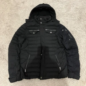 Svart quiltad jacka med huva - Snygg svart quiltad jacka från Performance med avtagbar huva och flera praktiska fickor med dragkedjor. Jackan har en stilren design med knappar och dragkedja framtill, perfekt för kyligare dagar. Midjan går att dras åt. Väldigt bra skick på jackan och nypriset var ungefär 1200 kr. Hör gärna av er om ni har frågor.