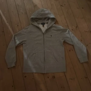Grå hoodie från Polo Ralph Lauren - Grå hoodie från Polo Ralph Lauren i mjuk bomullsblandning. Klassisk modell med dragkedja, huva och två fickor framtill. Liten broderad logga på bröstet och ribbade muddar vid ärmslut och nederkant. Perfekt för chill dagar.