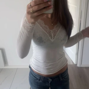 Vit spetsblus med v-ringning - Säljer en superfin vit blus med v-ringning och spetsdetaljer vid halsringning och ärmslut. Toppen är figurnära och har långa ärmar, perfekt att styla med jeans eller kjol. Materialet är mjukt och stretchigt för en bekväm passform.