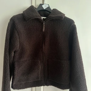 Mörkbrun teddyjacka med dragkedja - Mysig mörkbrun teddyjacka från H&M med hög krage och hel dragkedja framtill. Jackan har två stora fickor och är tillverkad i fluffigt fleece-material som håller dig varm under kyliga dagar. Perfekt för lager-på-lager och chill vardagsstil.