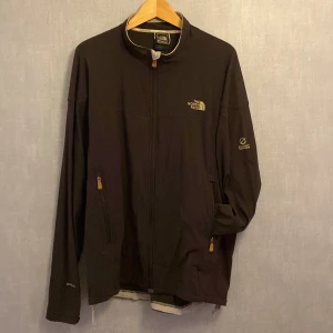 Svart vindjacka från The North Face XL - Svart vindjacka från The North Face Flight Series i storlek XL. Jackan har dragkedja framtill, två sidofickor med dragkedja och diskreta loggor på bröstet och ärmen. Tillverkad i polyester och perfekt för blåsiga dagar.