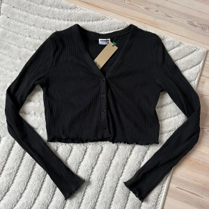 Svart croppad kofta från Noisy May - Svart ribbad kofta från Noisy May med croppad passform och v-ringning. Långa ärmar och knappar framtill. 
