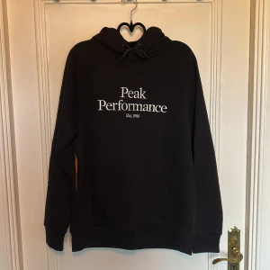 Svart hoodie från Peak Performance - Svart hoodie från Peak Performance med vit logga framtill. Klassisk modell med huva och snörning samt en stor magficka. Perfekt för dig som gillar en enkel och stilren look. Mjuk och skön, passar till många tillfällen.