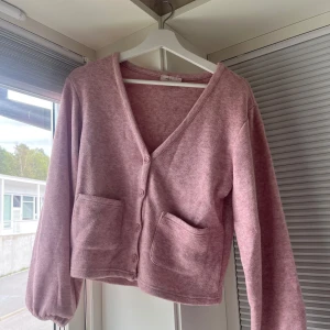 Rosa kofta från Nelly med fickor - Mysig rosa kofta från Nelly med v-ringning, knappar framtill och två stora fickor. Koftan har långärmad passform och är tillverkad i ett mjukt material som känns skönt mot huden. Perfekt för lager-på-lager och chill dagar.